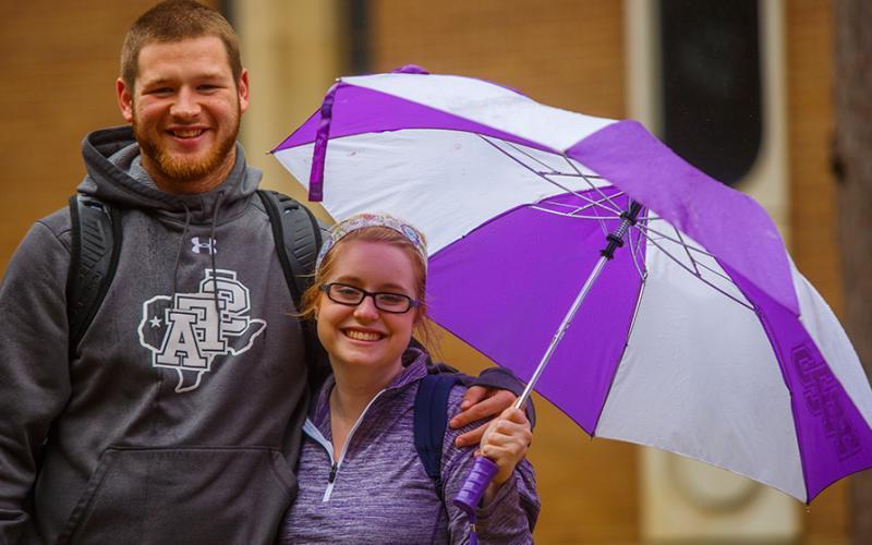 两名身着国家林书局服装的研究生站在一起，打着紫色和白色的雨伞
